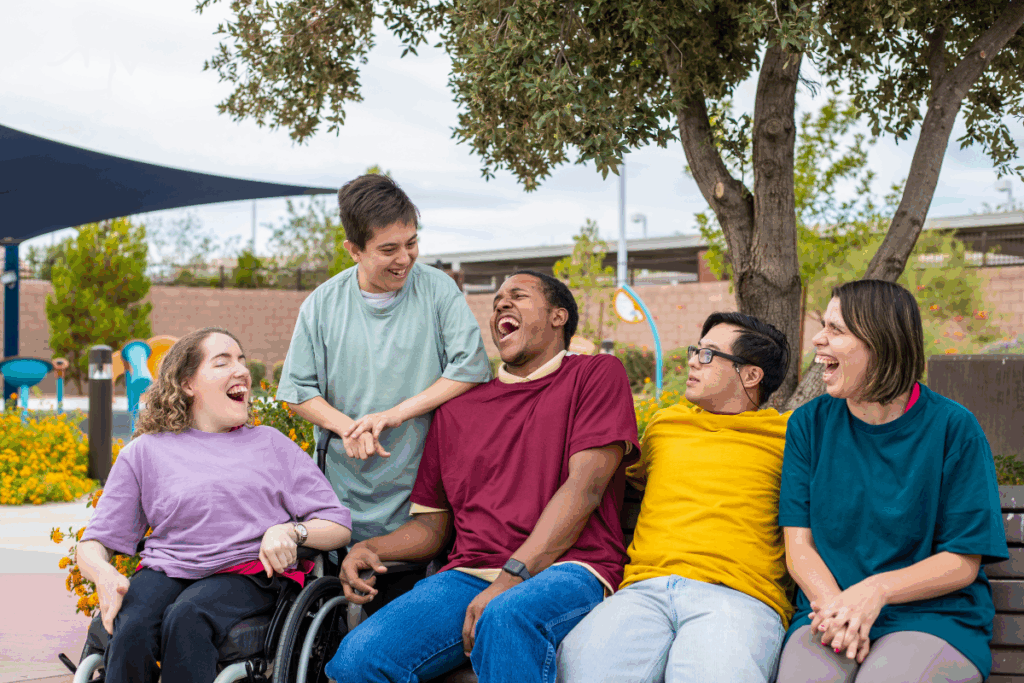 Groupe de jeunes adultes, dont une personne en fauteuil roulant, riant ensemble sur un banc dans un parc.